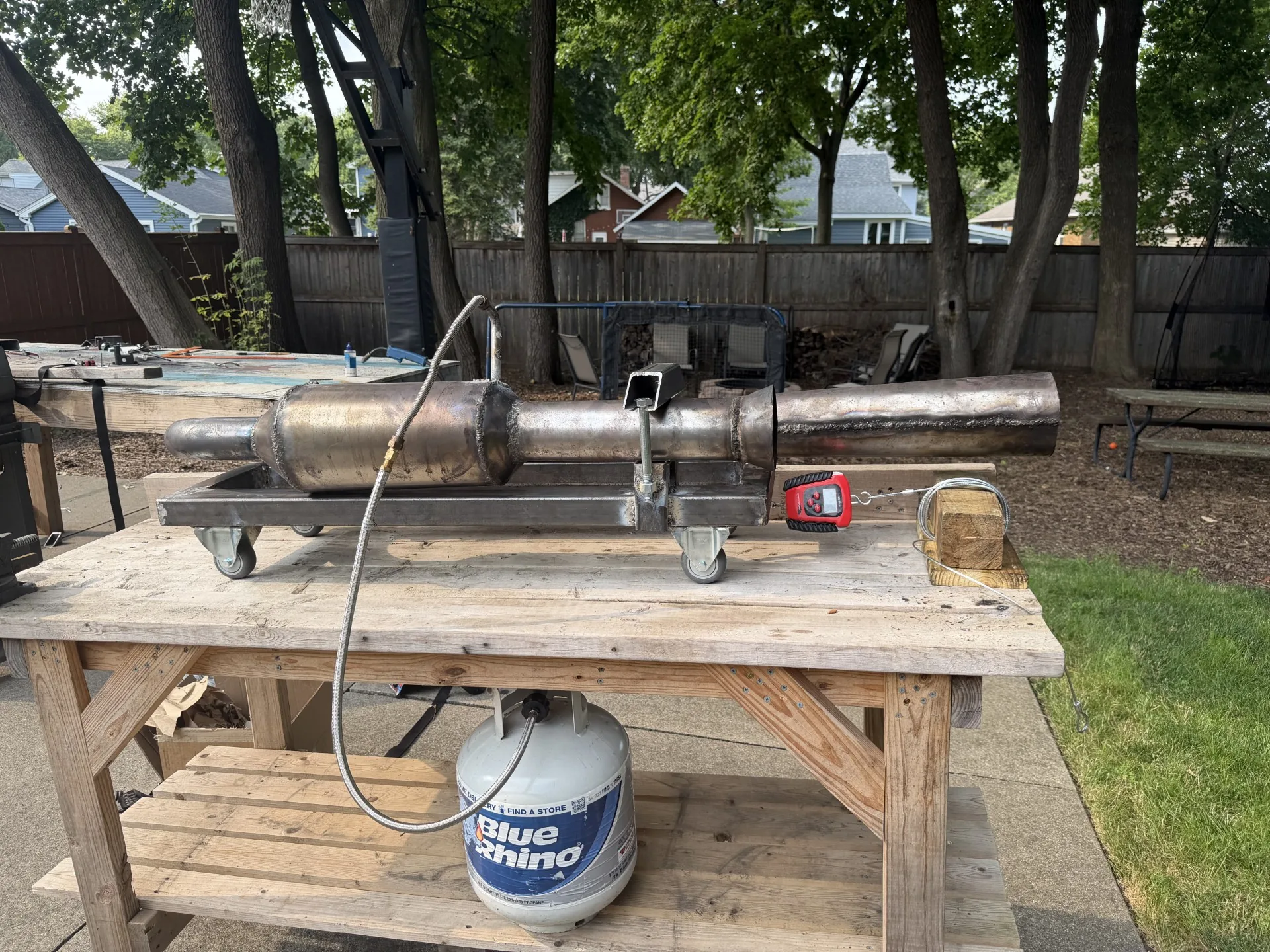 Steel pulse jet mounted on a wheeled frame on a wooden work table, with a propane tank on the shelf below, a braided fuel line, and a red digital scale tied to the frame